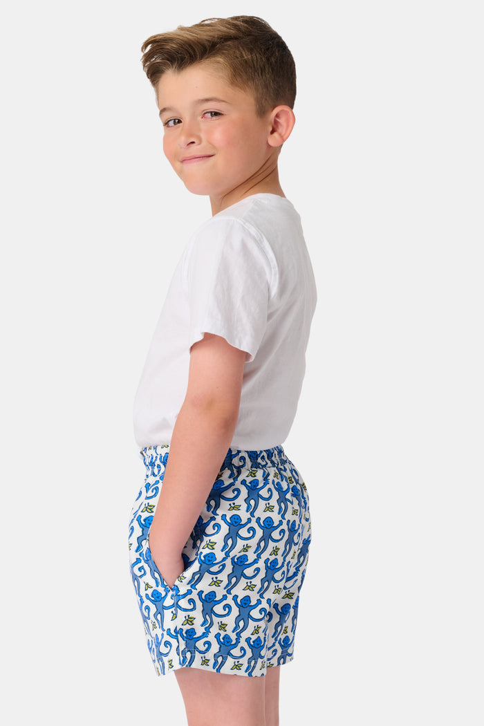 A child stands barefoot in a white t-shirt and Roller Rabbit Boys Monkey Swim Trunks, featuring a playful blue monkey print, with hands in pockets against a plain white background.