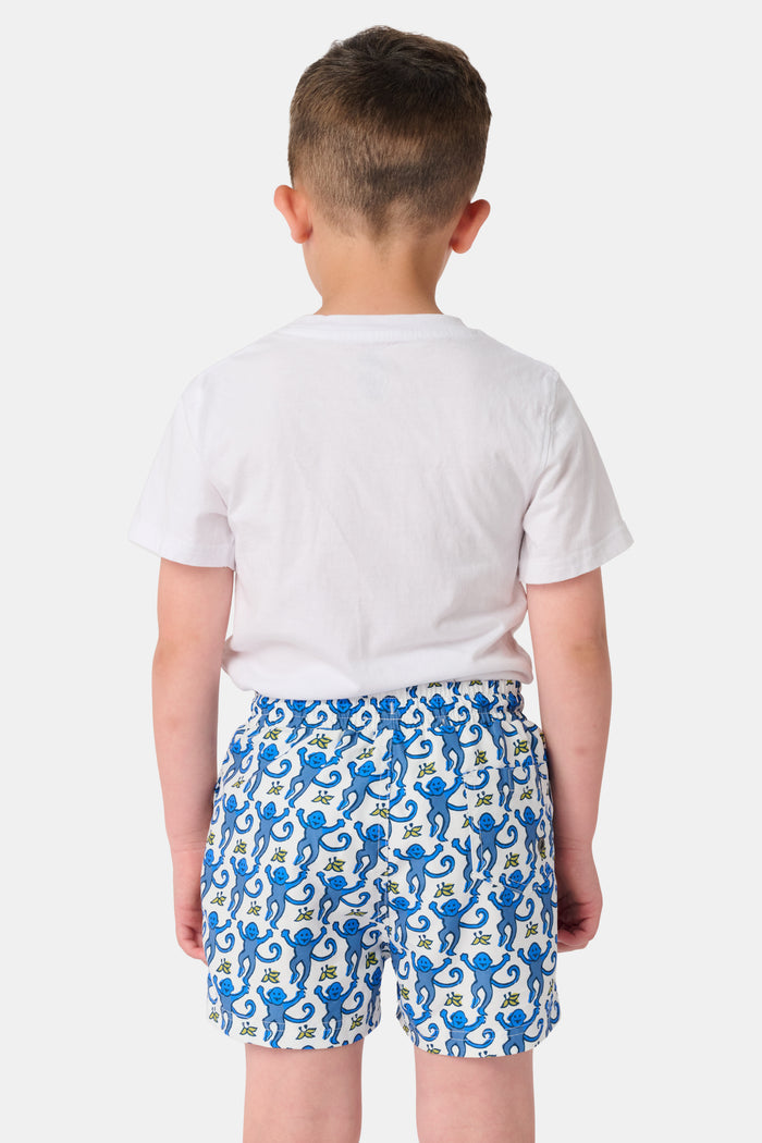 A child stands barefoot in a white t-shirt and Roller Rabbit Boys Monkey Swim Trunks, featuring a playful blue monkey print, with hands in pockets against a plain white background.
