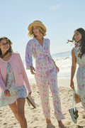 Three women smile as they walk barefoot on the beach, wearing colorful outfits. One wears the Roller Rabbit Camellia Block Millie Set. One holds sandals, another wears a straw hat, and the sparkling ocean is behind them.