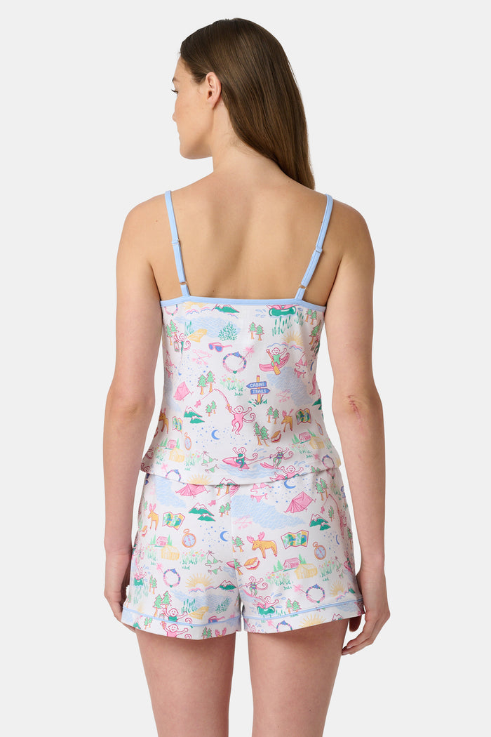 A woman with long brown hair wears the Roller Rabbit Camp RR Mischa Set, a light-hued Pima cotton pajama set with playful prints. The adjustable strap tank and shorts provide comfort as she stands turned slightly to the side, gazing off-camera.