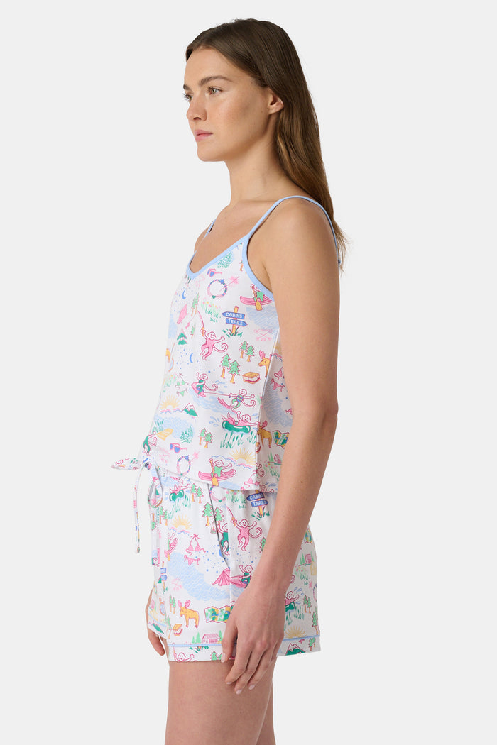 A woman with long brown hair wears the Roller Rabbit Camp RR Mischa Set, a light-hued Pima cotton pajama set with playful prints. The adjustable strap tank and shorts provide comfort as she stands turned slightly to the side, gazing off-camera.