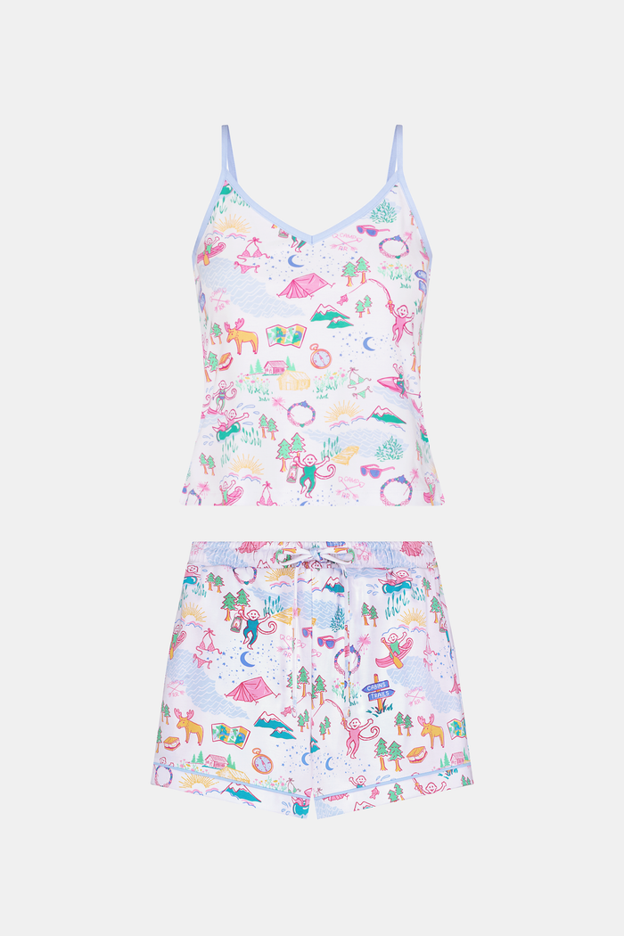 A woman with long brown hair wears the Roller Rabbit Camp RR Mischa Set, a light-hued Pima cotton pajama set with playful prints. The adjustable strap tank and shorts provide comfort as she stands turned slightly to the side, gazing off-camera.