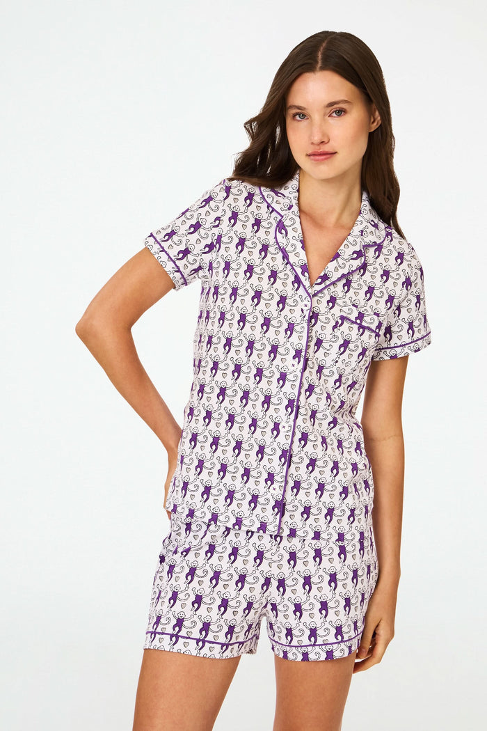 A woman with long brown hair wears the Roller Rabbit Campus Lovely Monkey Polo Pajamas—women's Pima cotton short-sleeve set with playful monkey print and purple trim—standing against a plain white background.