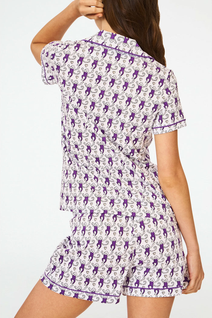 A woman with long brown hair wears the Roller Rabbit Campus Lovely Monkey Polo Pajamas—women's Pima cotton short-sleeve set with playful monkey print and purple trim—standing against a plain white background.
