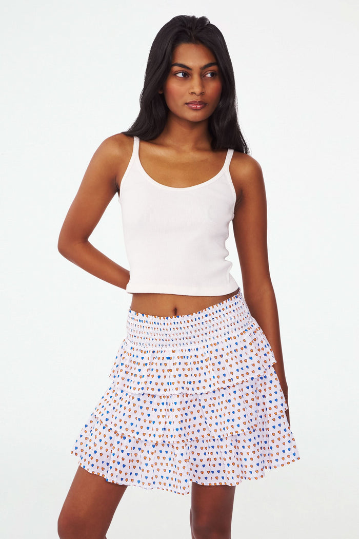 A person wearing the Roller Rabbit Campus Pop Hearts Siggy Skirt—a tiered ruffle mini with a smocked waist—and a white sleeveless ribbed top, stands with one hand on their hip against a white background.