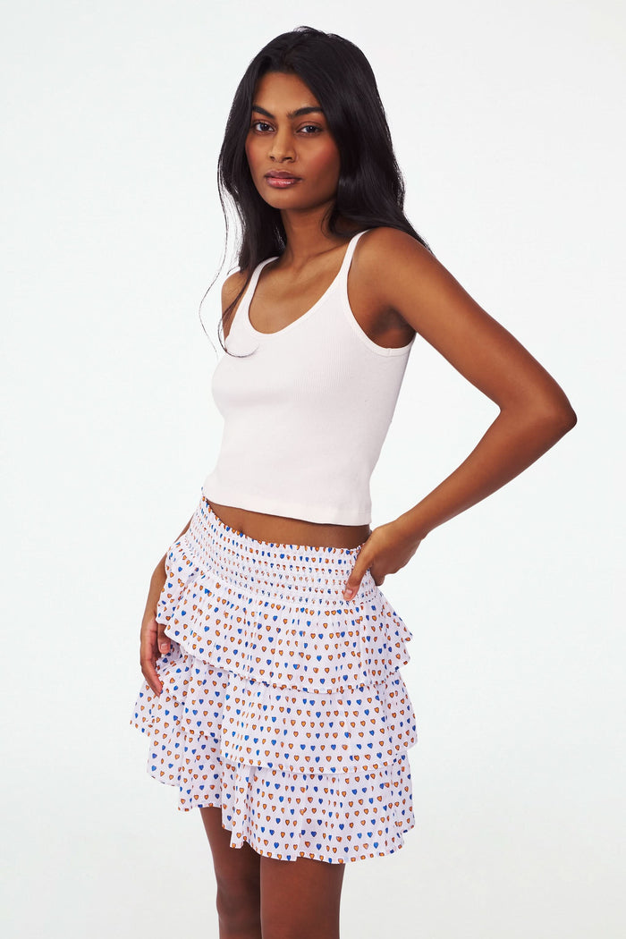 A person wearing the Roller Rabbit Campus Pop Hearts Siggy Skirt—a tiered ruffle mini with a smocked waist—and a white sleeveless ribbed top, stands with one hand on their hip against a white background.
