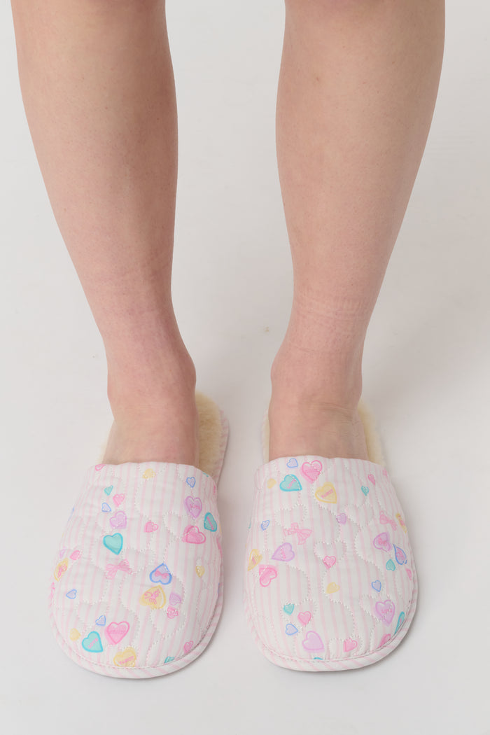A person stands on a white surface wearing Roller Rabbit Candy Heart Stripe Quilted Slippers. Only the lower legs and feet are visible.