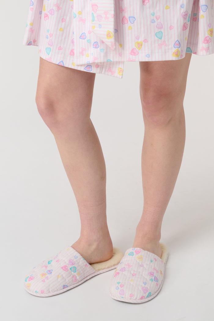 A person stands on a white surface wearing Roller Rabbit Candy Heart Stripe Quilted Slippers. Only the lower legs and feet are visible.
