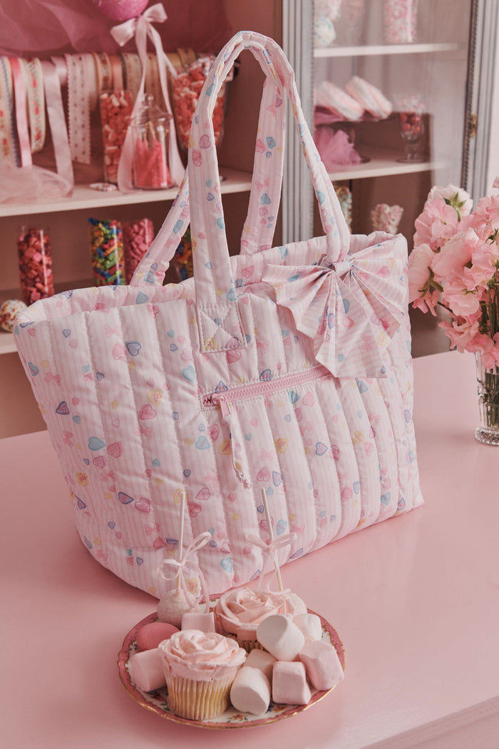 The Roller Rabbit Candy Heart Stripe Quilted Tote is a pastel pink weekend bag with scalloped edges, a matching bow on one handle, double handles, and a front zipper pocket.