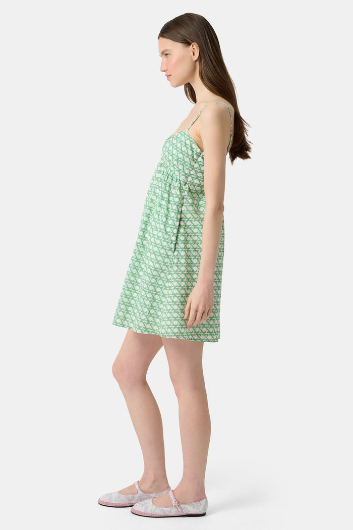 A woman with long curly hair wears the Roller Rabbit Cane Sofie Dress in kelly green, standing outdoors on grass by a hedge, with boats and water in the background.