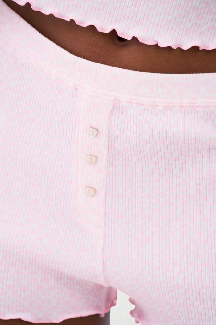A person wears the Roller Rabbit Catalina Rib Sullivan Shortie set—light pink ribbed tank top and matching boxer-inspired shorts with ruffled hems and decorative buttons—against a plain white background.