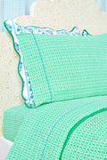 A close-up of a bed with green and white patterned Catalina Sheet Set from Roller Rabbit and a matching pillow with blue floral trim. The pale beige headboard features a floral pattern, adding style to this cozy 300-thread-count cotton bedding.