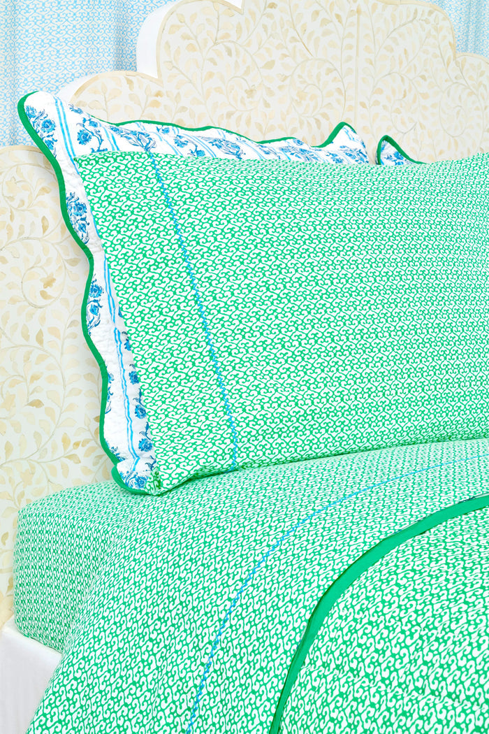 A close-up of a bed with green and white patterned Catalina Sheet Set from Roller Rabbit and a matching pillow with blue floral trim. The pale beige headboard features a floral pattern, adding style to this cozy 300-thread-count cotton bedding.
