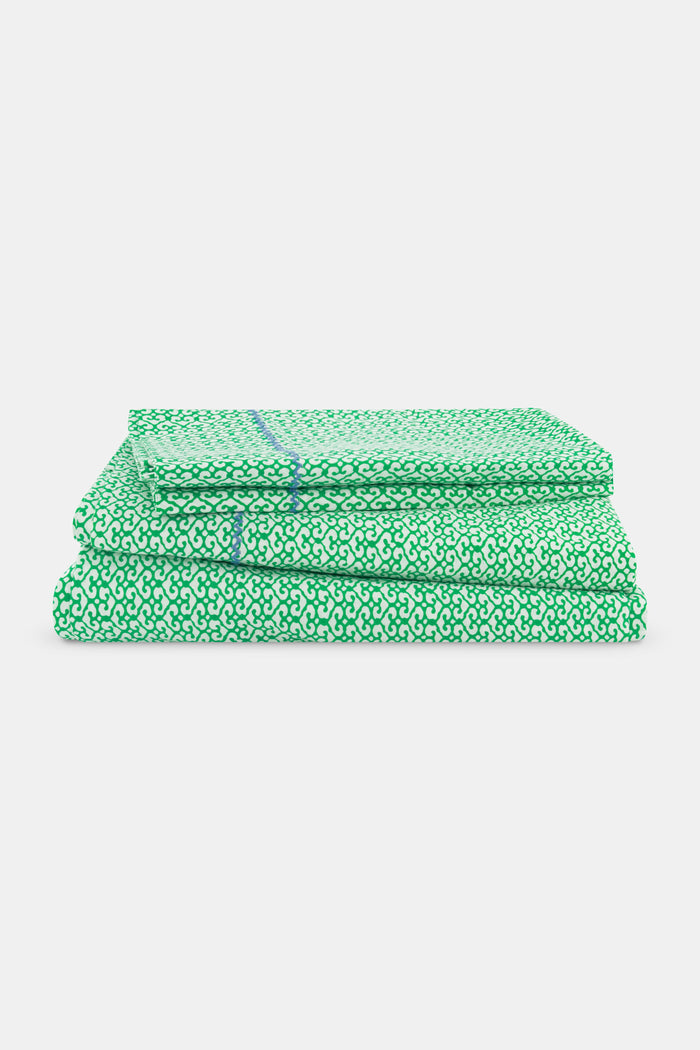 A close-up of a bed with green and white patterned Catalina Sheet Set from Roller Rabbit and a matching pillow with blue floral trim. The pale beige headboard features a floral pattern, adding style to this cozy 300-thread-count cotton bedding.