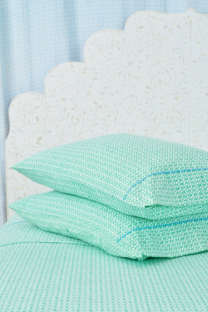 A close-up of a bed with green and white patterned Catalina Sheet Set from Roller Rabbit and a matching pillow with blue floral trim. The pale beige headboard features a floral pattern, adding style to this cozy 300-thread-count cotton bedding.