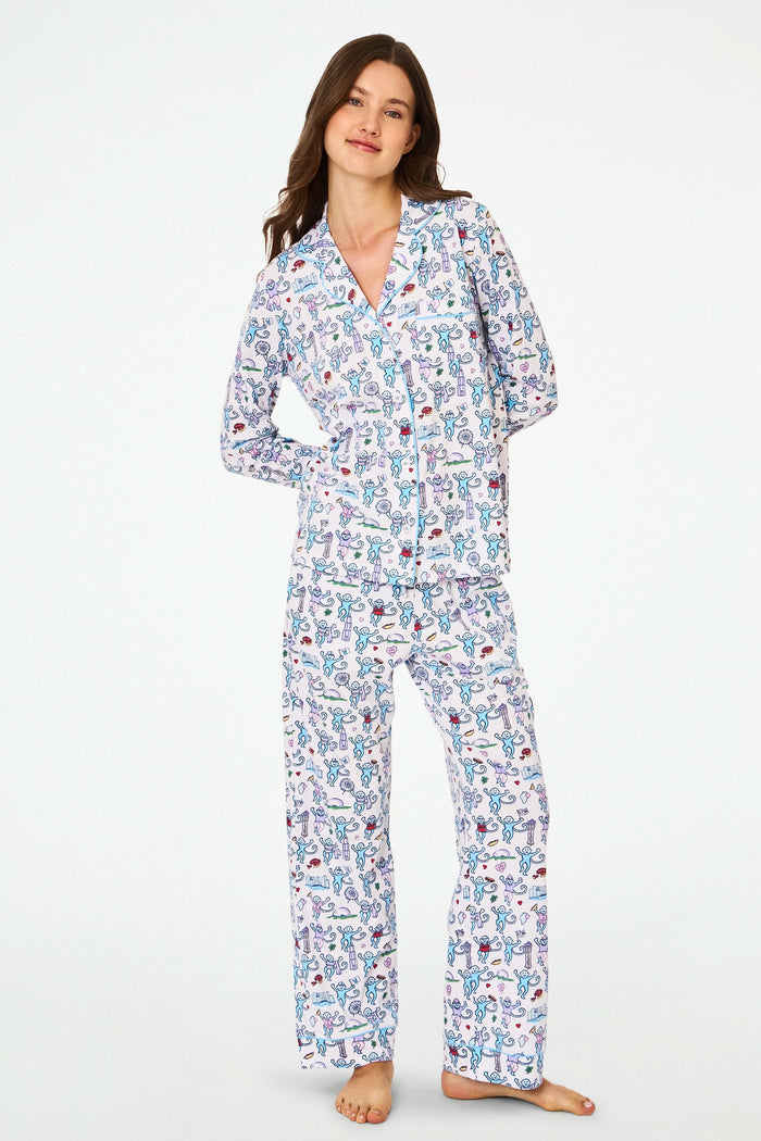 A woman smiles with her hands behind her back, barefoot against a white background, wearing Roller Rabbit’s Chicago Monkey Long Sleeve Polo Pajamas made from 100 Pima cotton with a vibrant, whimsical print.