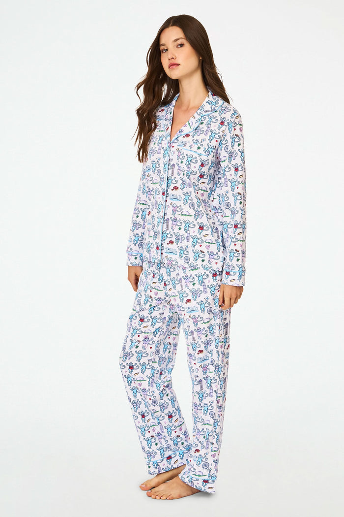 A woman smiles with her hands behind her back, barefoot against a white background, wearing Roller Rabbit’s Chicago Monkey Long Sleeve Polo Pajamas made from 100 Pima cotton with a vibrant, whimsical print.
