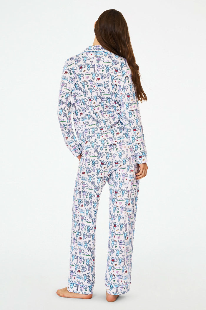 A woman smiles with her hands behind her back, barefoot against a white background, wearing Roller Rabbit’s Chicago Monkey Long Sleeve Polo Pajamas made from 100 Pima cotton with a vibrant, whimsical print.