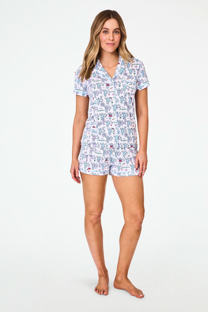 A woman smiles in Roller Rabbit's Chicago Monkey Polo Pajamas—a cozy Pima cotton set with a short-sleeved button-up top and shorts, featuring a playful, colorful monkey print on white—perfect for relaxed weekends.