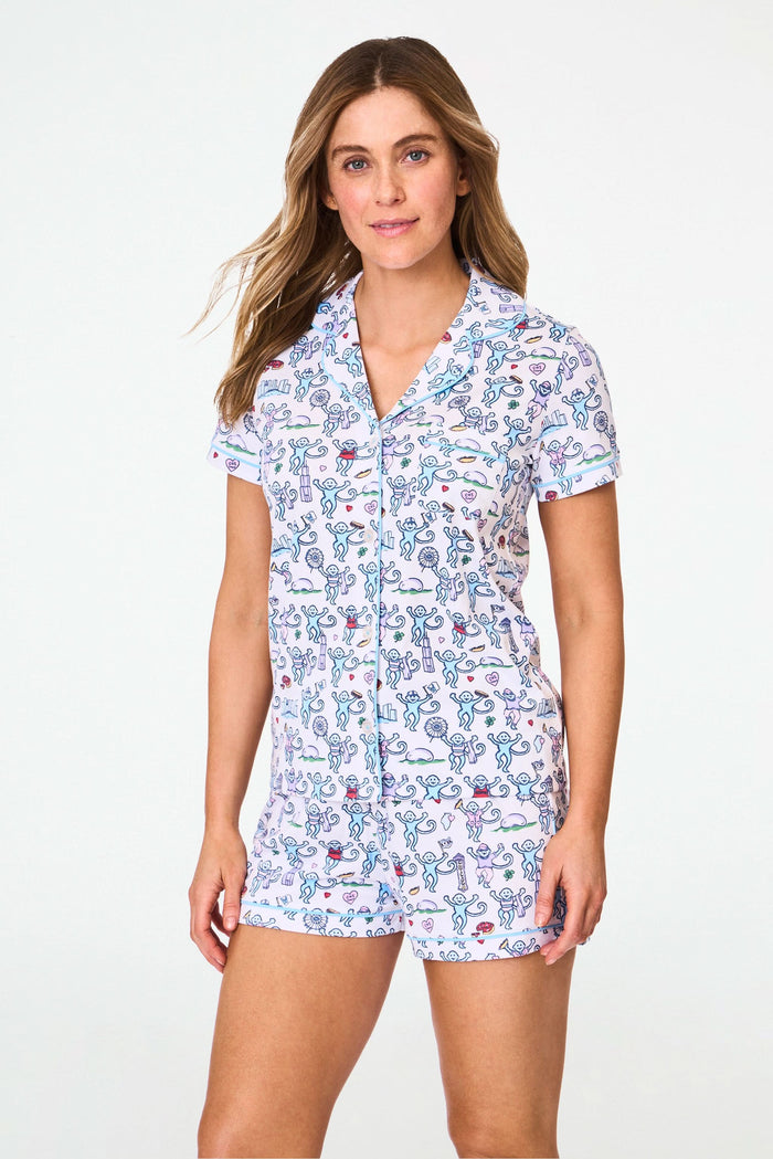 A woman smiles in Roller Rabbit's Chicago Monkey Polo Pajamas—a cozy Pima cotton set with a short-sleeved button-up top and shorts, featuring a playful, colorful monkey print on white—perfect for relaxed weekends.
