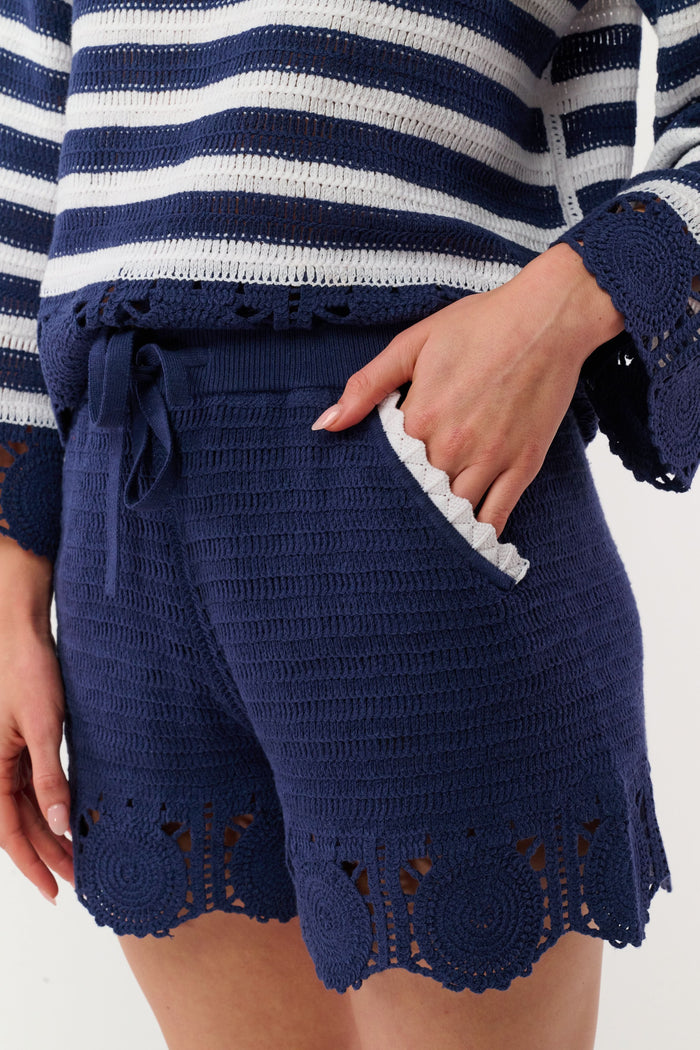 A person wears the Roller Rabbit Crochet Cameron Shorts—navy blue and white with an elastic waist, lace detailing, and a pocket—paired with a matching striped crochet sweater for chic beachwear.