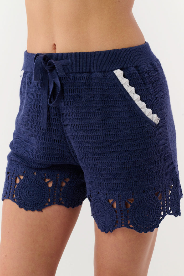 A person wears the Roller Rabbit Crochet Cameron Shorts—navy blue and white with an elastic waist, lace detailing, and a pocket—paired with a matching striped crochet sweater for chic beachwear.