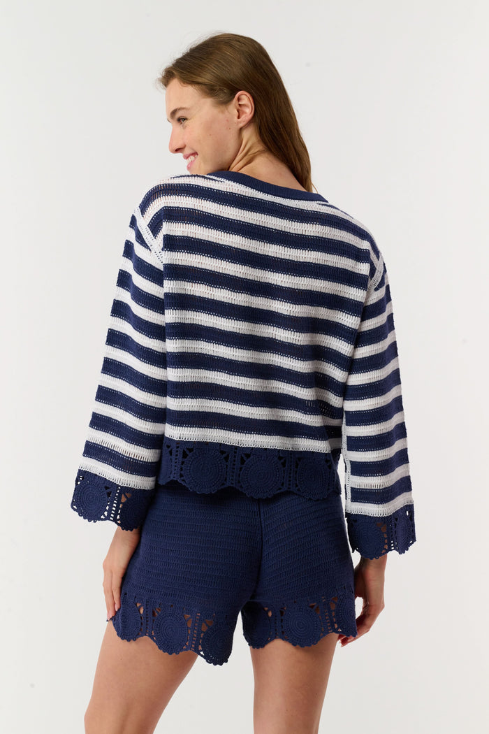A person wears the Roller Rabbit Crochet Cameron Shorts—navy blue and white with an elastic waist, lace detailing, and a pocket—paired with a matching striped crochet sweater for chic beachwear.