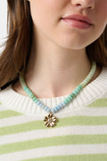 A person is shown from the shoulders up wearing a light green and white striped sweater and the Roller Rabbit Daisy Beaded Charm Necklace, which features pastel blue and green beads with a gold flower-shaped pendant.