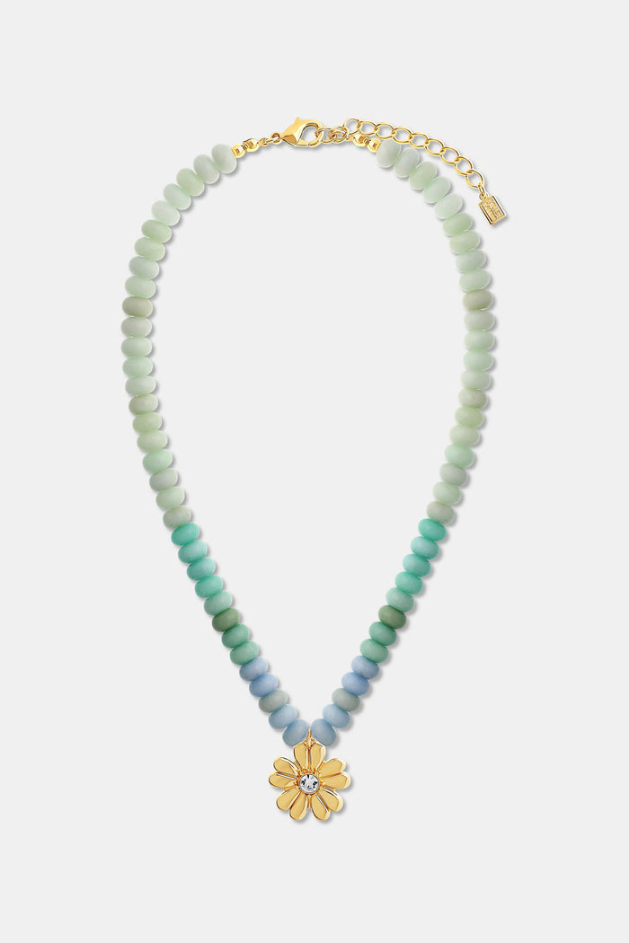 A person is shown from the shoulders up wearing a light green and white striped sweater and the Roller Rabbit Daisy Beaded Charm Necklace, which features pastel blue and green beads with a gold flower-shaped pendant.