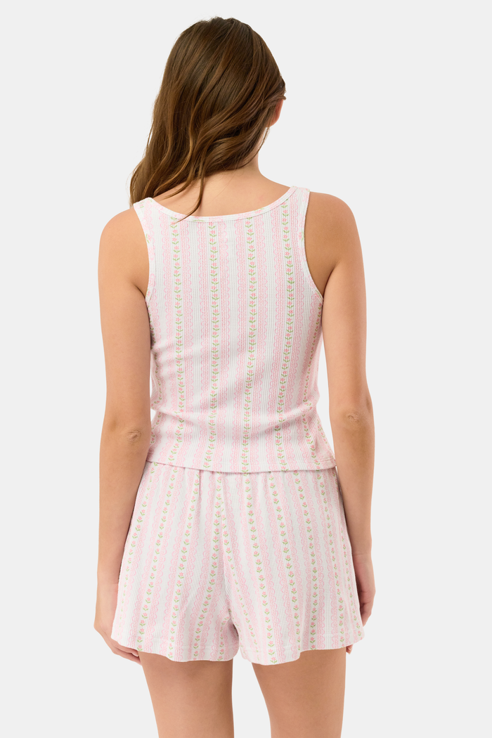 A woman stands barefoot against a white background, wearing the Roller Rabbit Daisy Heart Stripe Cherry Tank Set—a sleeveless, light pink loungewear set in Pima cotton with pastel vertical stripes, featuring a tank top and drawstring shorts.