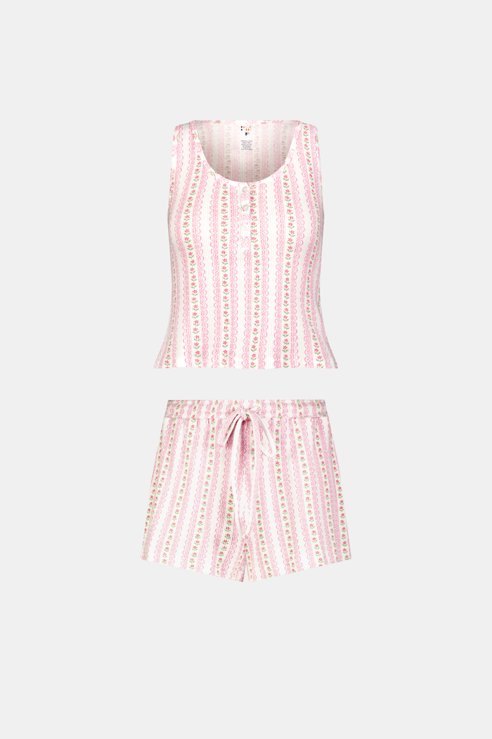 A woman stands barefoot against a white background, wearing the Roller Rabbit Daisy Heart Stripe Cherry Tank Set—a sleeveless, light pink loungewear set in Pima cotton with pastel vertical stripes, featuring a tank top and drawstring shorts.