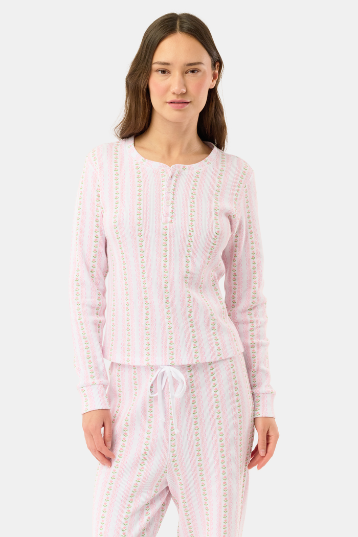 A woman wears the Roller Rabbit Daisy Heart Stripe Rosemary Top with pink, green, and white stripes, featuring a long-sleeved henley design, against a plain light background.