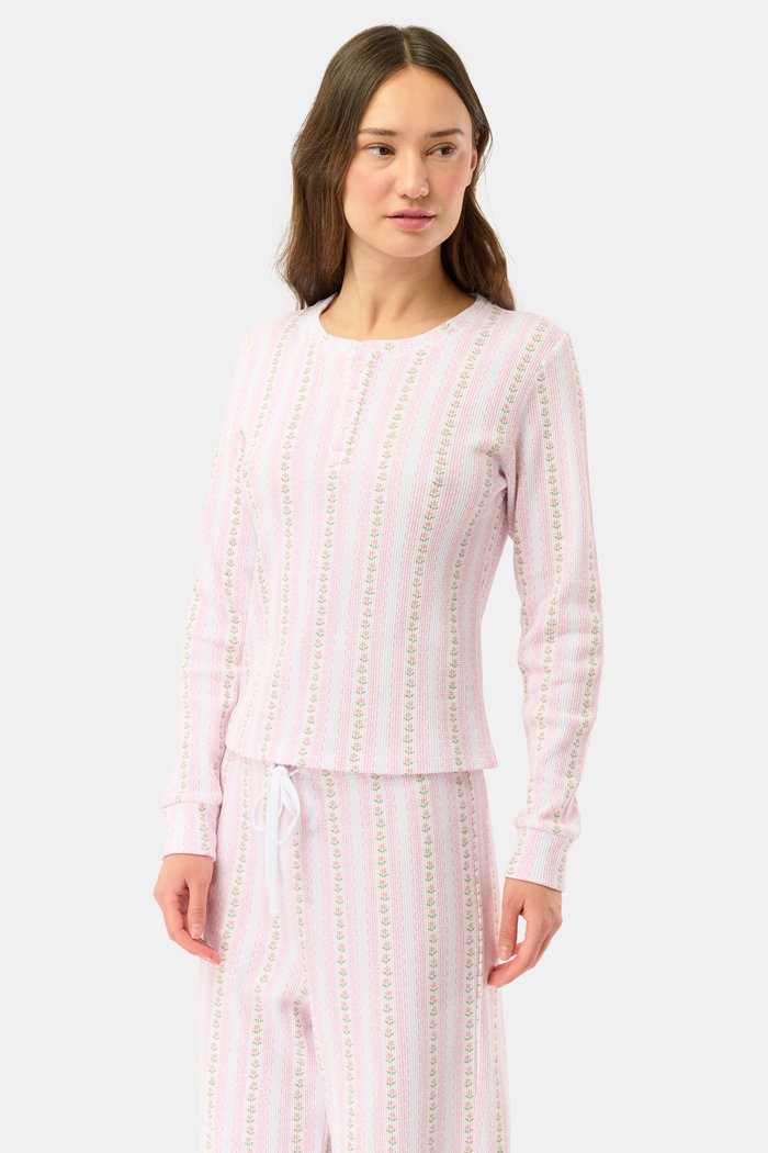 A woman wears the Roller Rabbit Daisy Heart Stripe Rosemary Top with pink, green, and white stripes, featuring a long-sleeved henley design, against a plain light background.