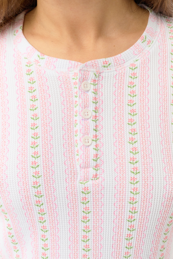 A woman wears the Roller Rabbit Daisy Heart Stripe Rosemary Top with pink, green, and white stripes, featuring a long-sleeved henley design, against a plain light background.