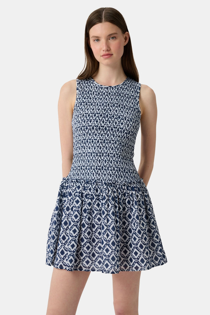 A woman with long straight hair and a neutral expression wears the Roller Rabbit Diamond Ikat Isla Dress—a navy smocked sleeveless dress with a white ikat pattern, flared skirt, and dropped waist, standing against a plain background.
