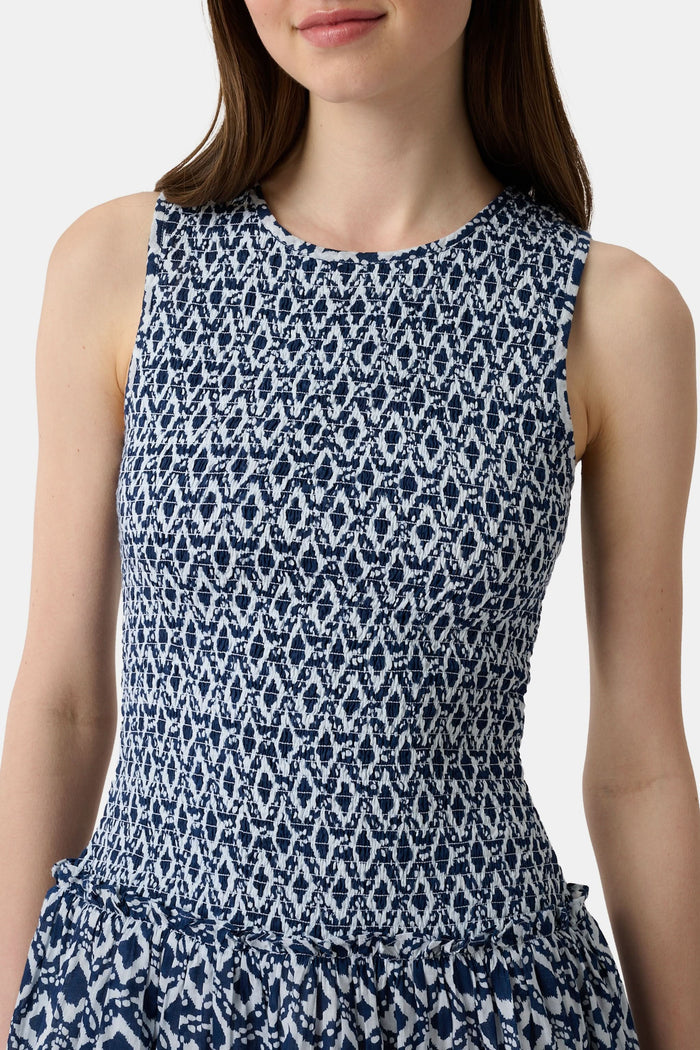 A woman with long straight hair and a neutral expression wears the Roller Rabbit Diamond Ikat Isla Dress—a navy smocked sleeveless dress with a white ikat pattern, flared skirt, and dropped waist, standing against a plain background.