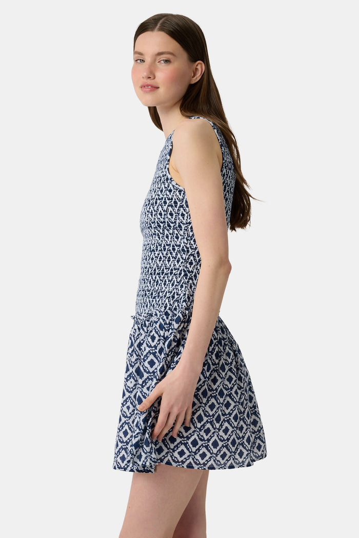 A woman with long straight hair and a neutral expression wears the Roller Rabbit Diamond Ikat Isla Dress—a navy smocked sleeveless dress with a white ikat pattern, flared skirt, and dropped waist, standing against a plain background.