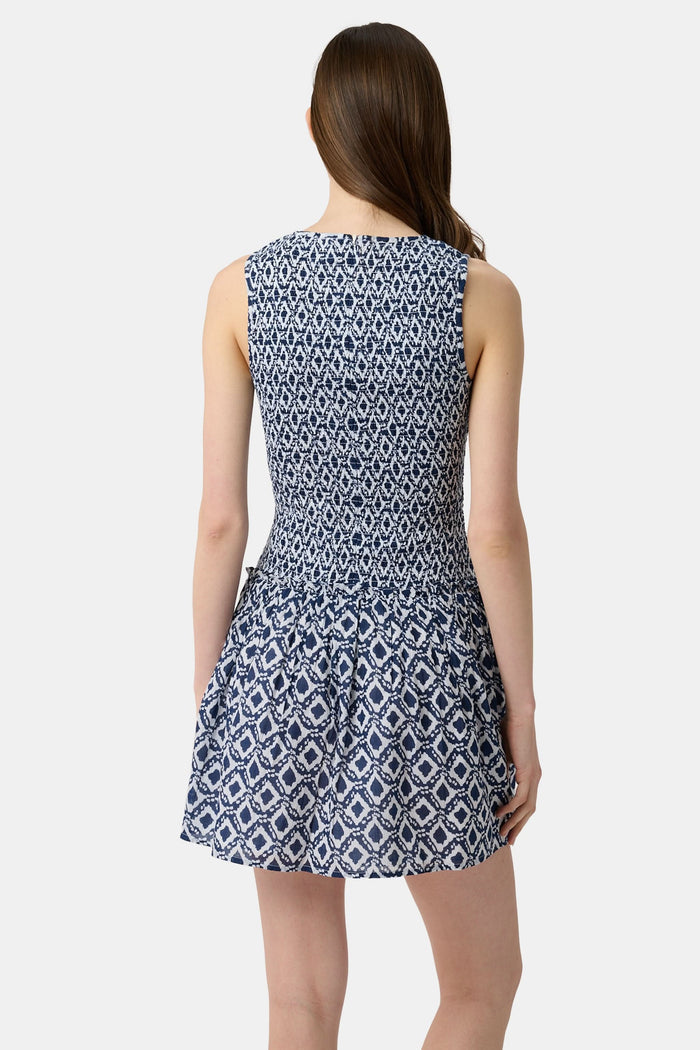 A woman with long straight hair and a neutral expression wears the Roller Rabbit Diamond Ikat Isla Dress—a navy smocked sleeveless dress with a white ikat pattern, flared skirt, and dropped waist, standing against a plain background.