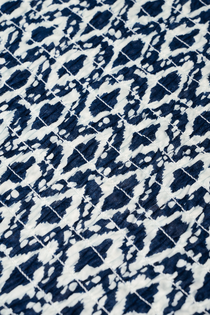 A woman with long straight hair and a neutral expression wears the Roller Rabbit Diamond Ikat Isla Dress—a navy smocked sleeveless dress with a white ikat pattern, flared skirt, and dropped waist, standing against a plain background.