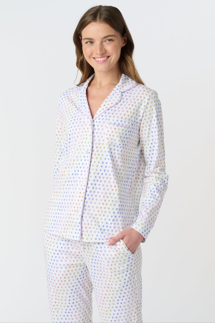 A woman stands barefoot against a plain background, wearing Roller Rabbit’s Disco Hearts Long Sleeve Polo Pajamas—a plush set made from 100 Pima cotton with a white base and small multicolored hearts, featuring long sleeves and front buttons.