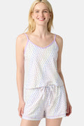 A woman smiles while wearing the Roller Rabbit Disco Hearts Mischa Set: a light-colored Pima cotton loungewear ensemble with a sleeveless, V-neck adjustable top and matching shorts, both adorned with small multicolored hearts, against a plain background.