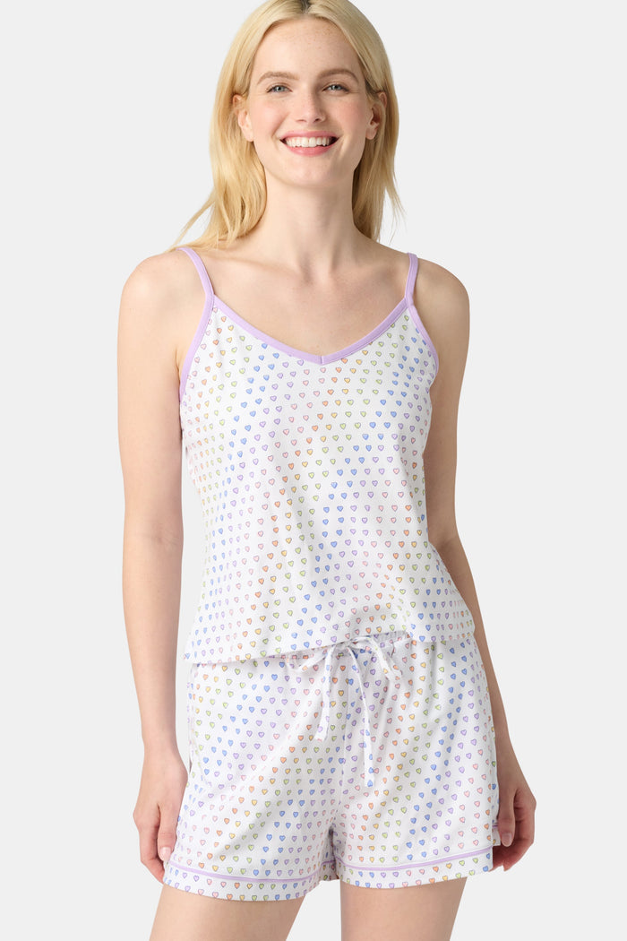 A woman smiles while wearing the Roller Rabbit Disco Hearts Mischa Set: a light-colored Pima cotton loungewear ensemble with a sleeveless, V-neck adjustable top and matching shorts, both adorned with small multicolored hearts, against a plain background.