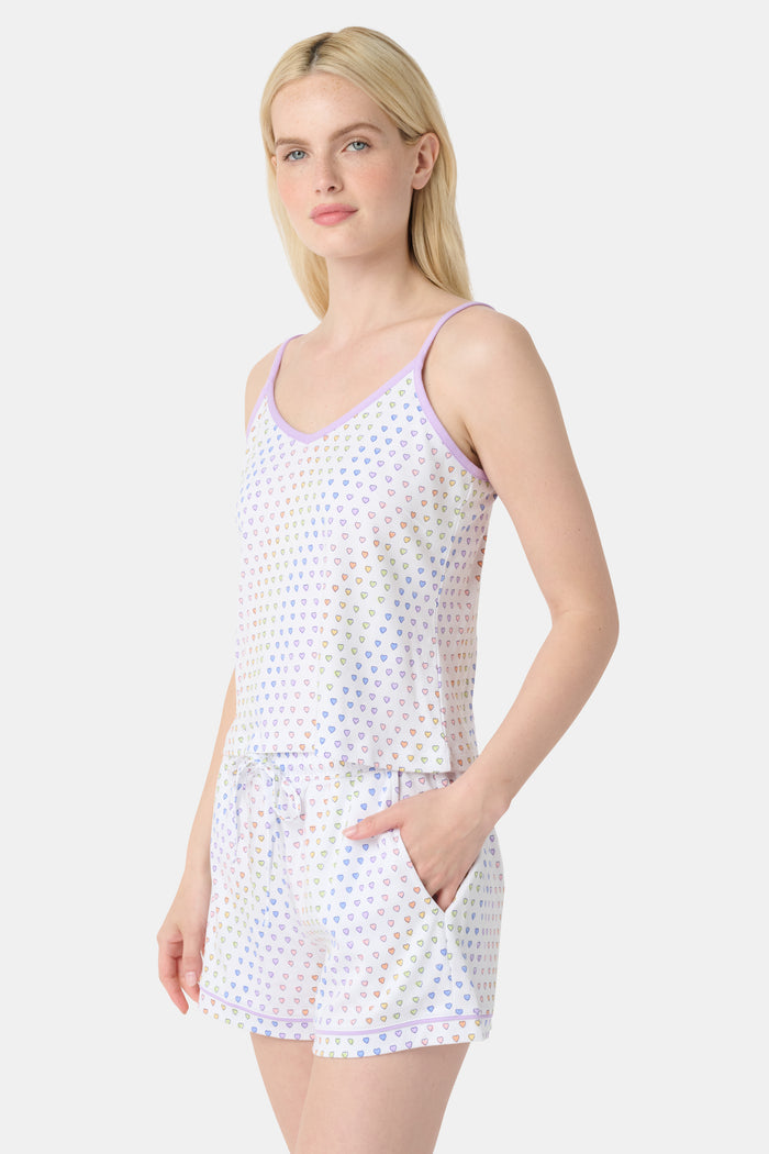 A woman smiles while wearing the Roller Rabbit Disco Hearts Mischa Set: a light-colored Pima cotton loungewear ensemble with a sleeveless, V-neck adjustable top and matching shorts, both adorned with small multicolored hearts, against a plain background.