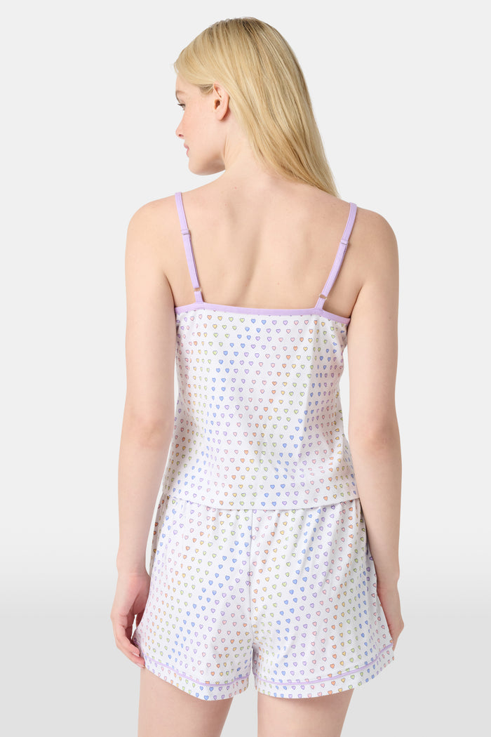A woman smiles while wearing the Roller Rabbit Disco Hearts Mischa Set: a light-colored Pima cotton loungewear ensemble with a sleeveless, V-neck adjustable top and matching shorts, both adorned with small multicolored hearts, against a plain background.
