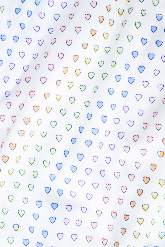 A woman smiles while wearing the Roller Rabbit Disco Hearts Mischa Set: a light-colored Pima cotton loungewear ensemble with a sleeveless, V-neck adjustable top and matching shorts, both adorned with small multicolored hearts, against a plain background.