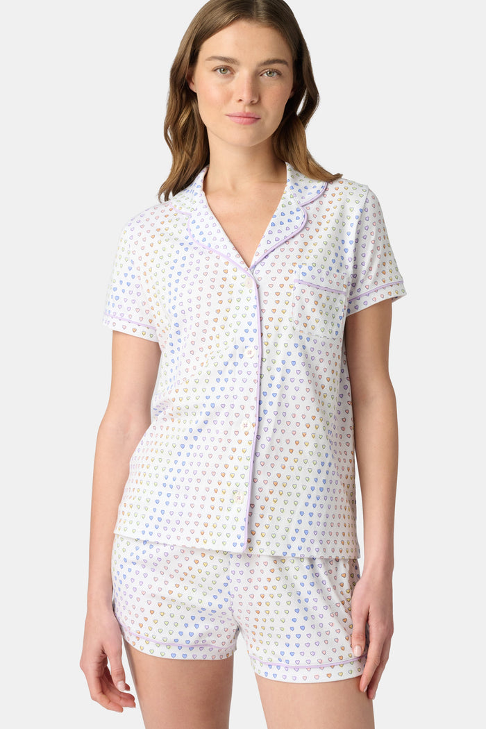 A woman wears the Roller Rabbit Disco Hearts Polo Pajamas, a white short-sleeve set with small multicolored hearts, standing against a plain light gray background.