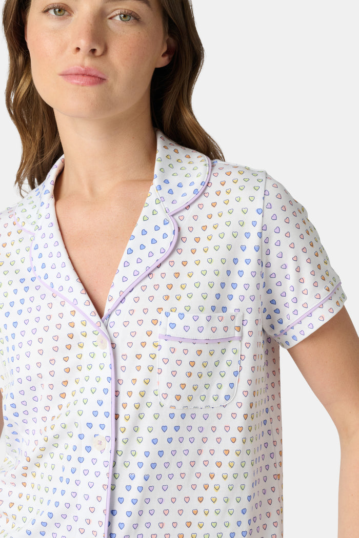 A woman wears the Roller Rabbit Disco Hearts Polo Pajamas, a white short-sleeve set with small multicolored hearts, standing against a plain light gray background.