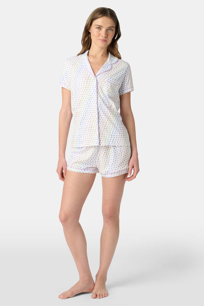 A woman wears the Roller Rabbit Disco Hearts Polo Pajamas, a white short-sleeve set with small multicolored hearts, standing against a plain light gray background.