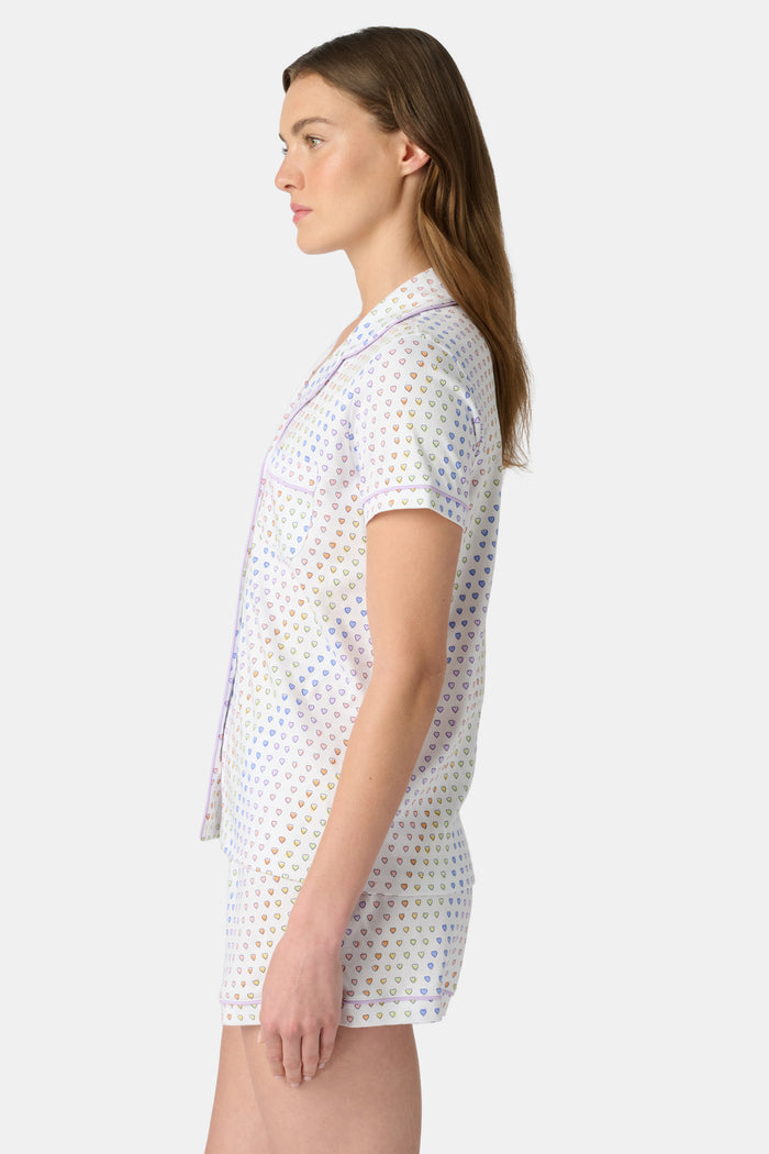 A woman wears the Roller Rabbit Disco Hearts Polo Pajamas, a white short-sleeve set with small multicolored hearts, standing against a plain light gray background.