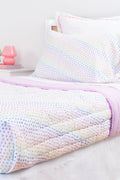 A neatly made twin bed features the Roller Rabbit Disco Hearts Quilt with white bedding and a colorful small heart pattern, complemented by a folded lavender blanket, pink lamp, and books on a white nightstand.